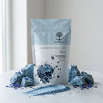 A stand-up pouch of Wild Organic Cambrian Blue Clay healing clay stands on a light surface with blue clay rocks, loose powder, and scattered blue flower petals around it. The pouch is pale blue and white with a printed tree logo and product name, photographed in a bright indoor setting.