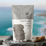 A stand-up pouch of Wild Organic Dead Sea Mud mineral rich clay, displayed on a dark stone slab with a wooden scoop of the gray mud and sea salt crystals, set against a blurred background of a blue ocean and sandy shore.