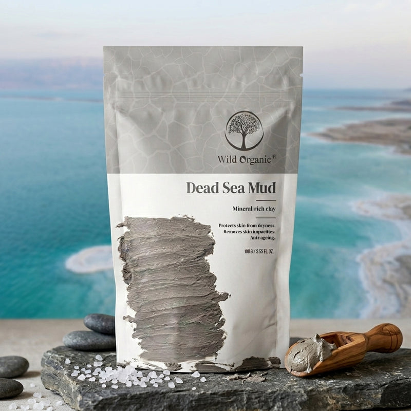 A stand-up pouch of Wild Organic Dead Sea Mud mineral rich clay, displayed on a dark stone slab with a wooden scoop of the gray mud and sea salt crystals, set against a blurred background of a blue ocean and sandy shore.