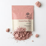 A stand-up pouch of Wild Organic French Pink Clay powder on a white background, with loose pink clay scattered around and in front of the package.