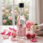 A bottle of Wild Organic Rose Water Skin Toner stands on a marble surface surrounded by pink rose petals, fresh roses, and a rolled towel, with a softly blurred window of pink flowers in the background.