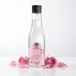 A clear plastic bottle of Wild Organic Rose Water San Tower with a black cap and a pink label, styled with pink rose petals and two pink roses on a white reflective surface against a white background.