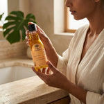 A woman in a cream robe holds a bottle of Wild Organic Laca de Oro hand and body lotion over a stone countertop in a warm, sunlit bathroom setting.