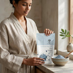 A woman in a beige linen robe holds a bag of Wild Organic Cambrian Blue Clay healing clay next to a ceramic bowl filled with the blue powder on a kitchen counter.