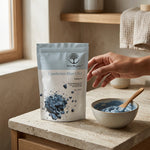 A stand-up pouch of Wild Organic Cambrian Blue Clay healing clay sits on a stone countertop beside a small bowl of blue clay and a wooden spoon, with a person's hand reaching toward the product in a warm, natural indoor setting.