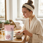 A woman in a beige robe and headband sits at a wooden table in a bright bathroom, mixing pink bath salts in a bowl. A pink and white pouch labeled Wild Organic French Pink Clay is placed on the table beside small bottles and folded towels.