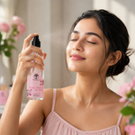 A woman with her eyes closed holds a clear spray bottle of Rose Water Skin Toner, spraying a fine mist. She is wearing a light pink camisole, and soft pink flowers are blurred in the background.